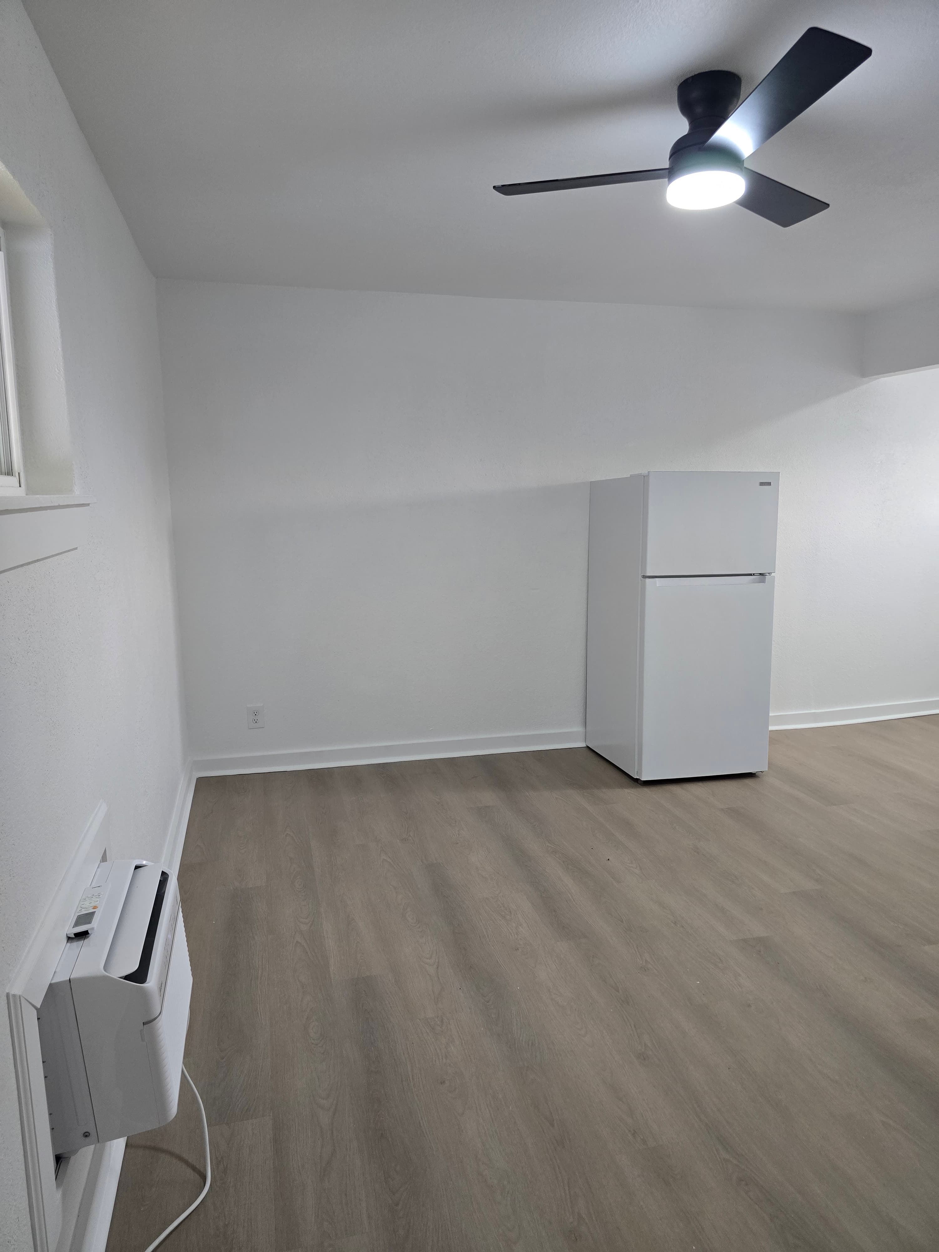 Renovated studio living area with new flooring, ceiling fan, and refrigerator